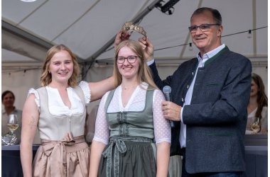 Bürgermeister Steinmacher im grauen Trachtenjacket und die ehemalige Weinkönigin in beige-weißem Dirndl halten eine goldene Krone mit Gemeindewappen, Reben und Trauben über den Kopf der neuen Weinkönigin im grün-weiss- farbenem Dirndl.