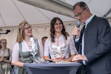 Zwei junge Frauen stehen hinter einem Bistrotisch. Rechts davon steht im grauem Trachten Jacket, Bürgermeister Steinmacher, mit Mikrofon in der Hand. Die linke Frau, die Weinkönigin trägt ein grün-weiss-farbenes Dirndl und eine goldene Krone mit Gemeindewappen, Reben und Trauben auf dem Kopf. In der Mitte die künftige Weinprinzessin hält ein Weinglas in der Hand und trägt ein grau-rosa-weißes Dirndl.