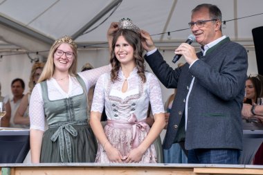 Bürgermeister Steinmacher im grauen Trachtenjacket und die Weinkönigin in grün-weiss-farbenem Dirndl halten eine silberne Krone mit Gemeindewappen, Reben und Trauben über den Kopf der neuen Weinpronzessin im rosa-weiß-farbenem Dirndl.  