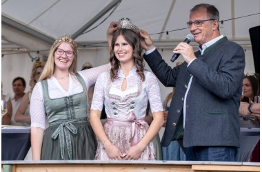Bürgermeister Steinmacher im grauen Trachtenjacket und die Weinkönigin in grün-weiss-farbenem Dirndl halten eine silberne Krone mit Gemeindewappen, Reben und Trauben über den Kopf der neuen Weinpronzessin im rosa-weiß-farbenem Dirndl.