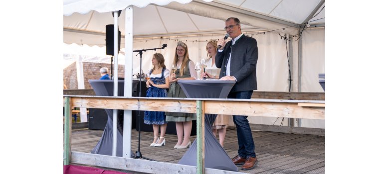 Drei junge Frauen stehen unter einem weißem Pavillon auf einer Holzbühne, hinter zwei Bistrotischen. Rechts davon steht im grauem Trachten Jacket, Bürgermeister Steinmacher, mit Mikrofon in der Hand. Die linke Frau, die Weinprinzessin trägt ein blau-weißes Dirndl. In der Mitte, die künftige Weinkönigin trägt ein weiß-grünes Dirndl und rechts die ehemalige Weinköniging, trägt ein weiß-grau-rosanes Dirndl. Alle haben ein Weinglas in der Hand und eine Krone auf dem Kopf.