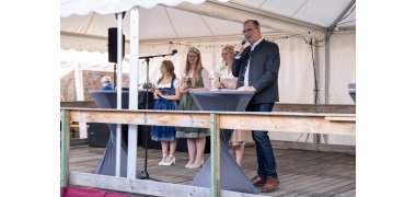 Drei junge Frauen stehen unter einem weißem Pavillon auf einer Holzbühne, hinter zwei Bistrotischen. Rechts davon steht im grauem Trachten Jacket, Bürgermeister Steinmacher, mit Mikrofon in der Hand. Die linke Frau, die Weinprinzessin trägt ein blau-weißes Dirndl. In der Mitte, die künftige Weinkönigin trägt ein weiß-grünes Dirndl und rechts die ehemalige Weinköniging, trägt ein weiß-grau-rosanes Dirndl. Alle haben ein Weinglas in der Hand und eine Krone auf dem Kopf.