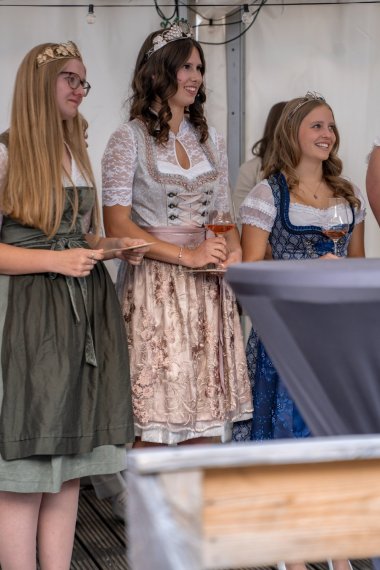 Drei junge Frauen stehen hinter einem Bistrotisch. Links die Weinkönigin trägt ein grün-weiss-farbenes Dirndl und eine goldene Krone mit Gemeindewappen, Reben und Trauben auf dem Kopf. In der Mitte steht die Weinprinzessin und hält ein Weinglas in der Hand, hat eine silberne Krone auf dem Kopf und ein grau-rosa-weißes Dirndl an. Rechts davon steht die Weinprinzessin im blau-grau-weiß-farbenem Dirndl mit silberner Krone auf dem Kopf und mit Rosé gefülltem Weinglas in der Hand.