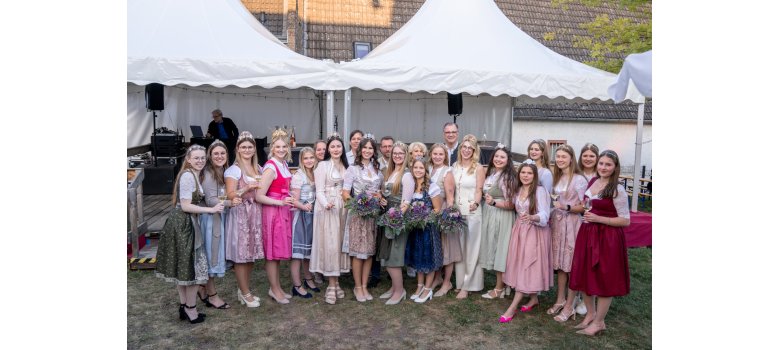 Vor einem großen, weißen Doppelpavillon stehen 17 Weinmajestäten in verschieden farbigen Dirndeln mit Krone auf dem Kopf und Weinglas in den Händen im Halbkreis. Drei Damen in der Mitte haben je einen Blumenstrauß in der Hand. Im Hintergrund des Halbkreises sind weitere Männer und Frauen. 