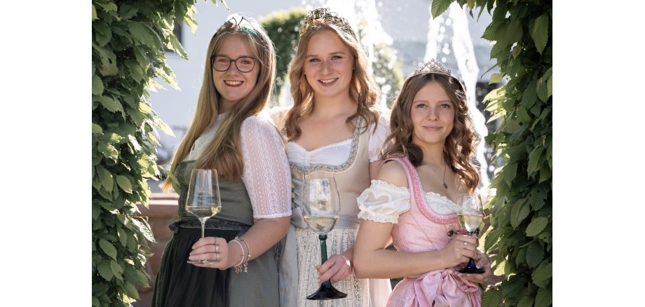 Die drei Kiedricher Weinmajestäten stehen nebeneinander. Alle haben ein gefülltes Weinglas in der Hand. Sie tragen ein rosa, ein beiges und ein grünes Dirndl, jeweils mit weißen Blusen. Sie tragen die blonden Haare offen, zwei Frisuren sind etwas gelockt. Die Dame mit den glatten Haaren trägt eine dunkle, runde Brille. Alle tragen ihre Krone auf dem Kopf. Umrandet ist das Bild von grünen Blättern.