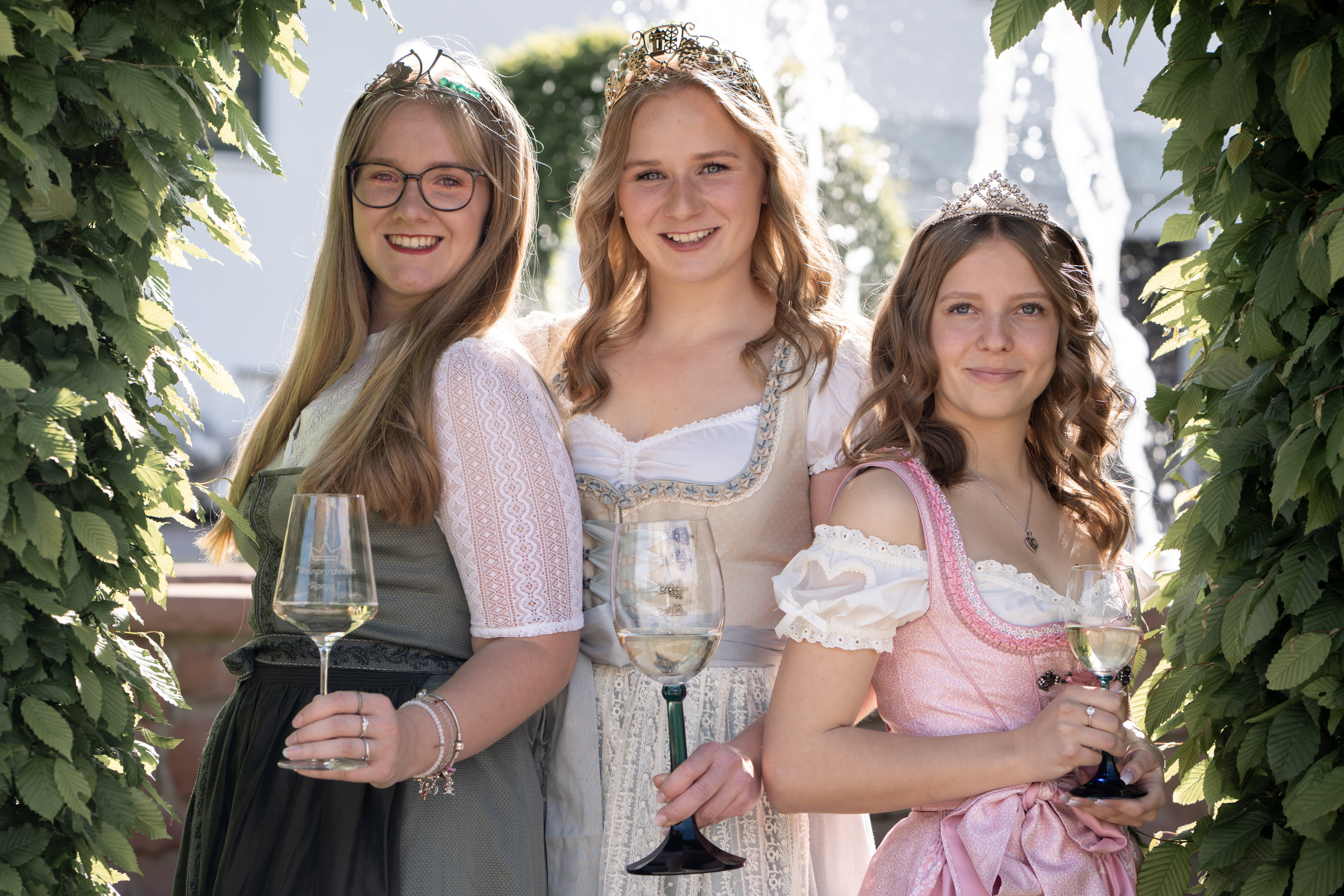 Die drei Kiedricher Weinmajestäten stehen nebeneinander. Alle haben ein gefülltes Weinglas in der Hand. Sie tragen ein rosa, ein beiges und ein grünes Dirndl, jeweils mit weißen Blusen. Sie tragen die blonden Haare offen, zwei Frisuren sind etwas gelockt. Die Dame mit den glatten Haaren trägt eine dunkle, runde Brille. Alle tragen ihre Krone auf dem Kopf. Umrandet ist das Bild von grünen Blättern.