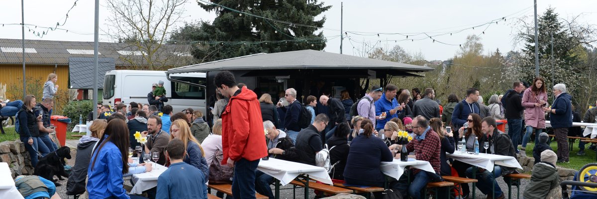 Viele Personen mit Jacken auf Bierzeltgarnituren auf einem Kiesboden vor dem Weinstand und Fahnenmasten mit Lichtergirlanden. 