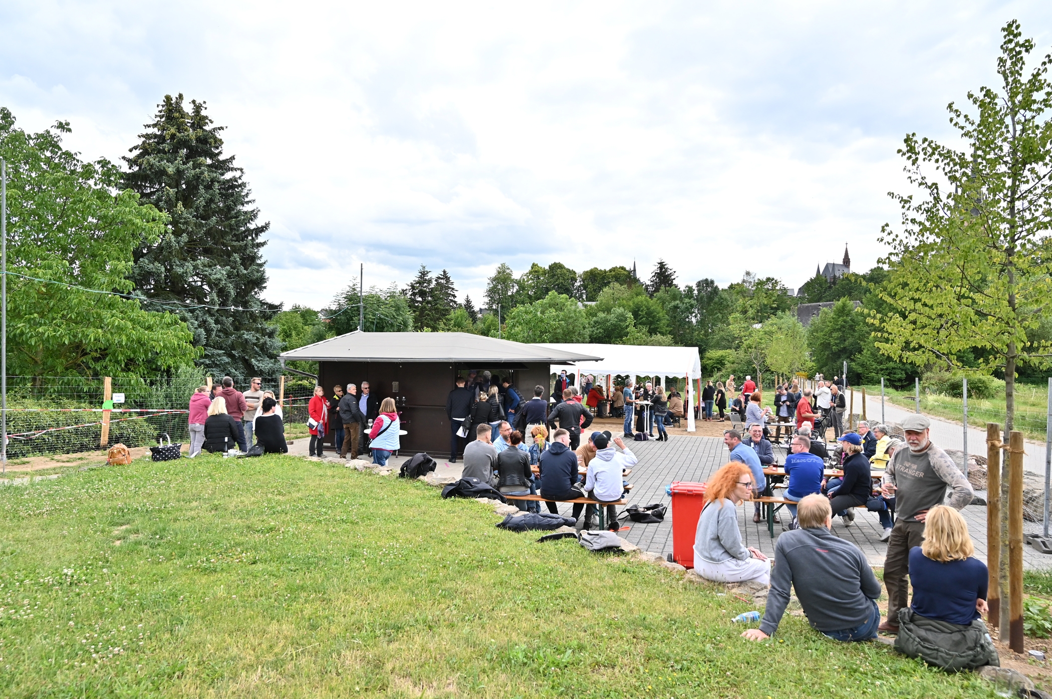 Mehrere Personen mit Jacken oder Pullovern auf Bierzeltgarnituren neben dem Weinstand. Ebenso ein weißes, rechteckiges Pavillon, asphaltierter, landwirtschaftlicher Weg, Rasen und im Hintergrund Bäume und der Kirchturm der Kirche St. Valentinus und Dionysius.