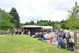 Mehrere Personen mit Jacken oder Pullovern auf Bierzeltgarnituren neben dem Weinstand. Ebenso ein weißes, rechteckiges Pavillon, asphaltierter, landwirtschaftlicher Weg, Rasen und im Hintergrund Bäume und der Kirchturm der Kirche St. Valentinus und Dionysius.