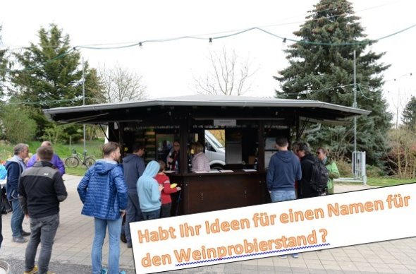 Mehrere Personen mit Jacken stehen an dem geöffneten, sechseckigen Weinstand aus Holz an. Ebenso sind Fahnenmasten mit Lichtergirlanden und Bäume erkennbar. Unten rechts steht eine Aufforderung der Nennung einer Idee für einen Namen für den Weinstand.