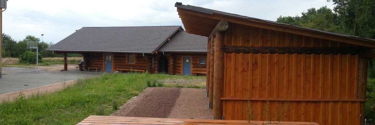Blick auf das Holzhaus mit blauen Türen und einem großen an einer Seite offeneer Holzuntersteller mit Kiesboden. Auf der Wiese steht eine aus Baumstämmen gefertigte Bank mit Tisch. Links von dem Blockhaus ist ein Basketballkorb aus Metall und ein aspahltierter Platz zu erkennen. Im Hintergrund sind belaubte Bäume. 