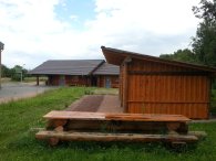 Jugendtreff "Easy" Blick auf das Holzhaus mit blauen Türen und einem großen an einer Seite offeneer Holzuntersteller mit Kiesboden. Auf der Wiese steht eine aus Baumstämmen gefertigte Bank mit Tisch. Links von dem Blockhaus ist ein Basketballkorb aus Metall und ein aspahltierter Platz zu erkennen. Im Hintergrund sind belaubte Bäume.