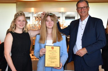 Die Kiedricher Weinprinzessin im schwarzen Kleid mit goldenem Diadem im Haar steht neben der Weinprinzessin mit langen blonden Haaren, weißem Shirt und hellblauem Anzug. Rechts davon steht Bürgermeister Winfried Steinmacher im weißem Hemd mit dunklen Knöpfen und einem dunkelblauem Anzug. Die Weinkönigin und der Bürgermeister setzen das Diadem der Weinprinzessin auf den Kopf, die eine beige Urkunde, die in einem goldenem Rahmen gerahmt ist, in den Händen hält.