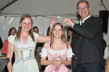Bürgermeister Steinmacher im schwarzen Anzug und die Weinkönigin in beige-weißem Dirndl halten eine Krone über den Kopf der neuen Weinprinzessin im rosa-weiss- farbenem Dirndl  