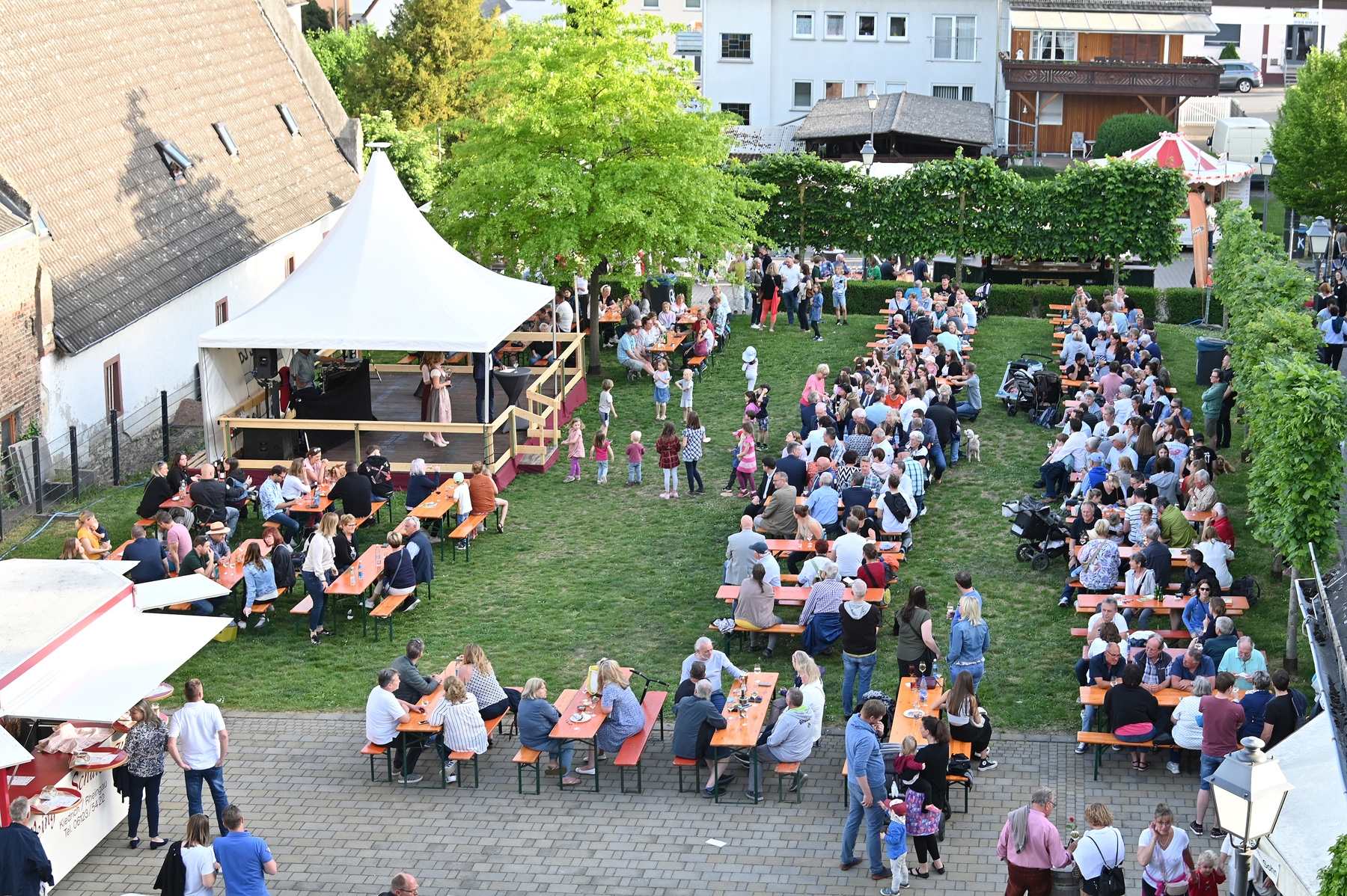 Blick auf eine Wiese, auf der eine Bühne mit einem weißem Dachzelt ist und viele Bierzeltgarnituren stehen, auf denen Menschen sitzen. Links im Vordergrund ist ein geöffneter Weinstand  zu sehen. Rechts von der Bühne ist ein grüner Laubbaum. An zwei Seiten des Festplatzes sind Reihen von schmalen Laubbäumen.