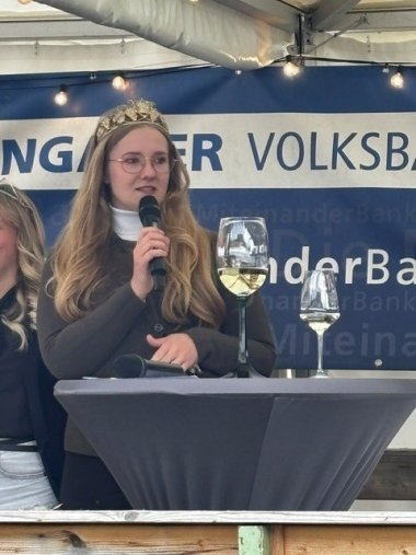 Eine Frau mit goldener Brille und goldener Krone aus Weinblättern und Reben, im langen blondem Haar, hält ein Mikrofon in der Hand. Sie trägt einen weißen Rollkragenpullover und eine olivgrüne Jacke. Sie steht hinter einem Bistrotisch mit einer grauen Husse, auf dem ein großes und ein kleines, jeweils gefülltes Weinglas stehen. Ebenso liegt ein Mikrofon auf dem Tisch. Im Hintergrund ist ein blau-weißes Banner zu sehen und das weiße Dach des Pavillons mit einer Lichterkette.