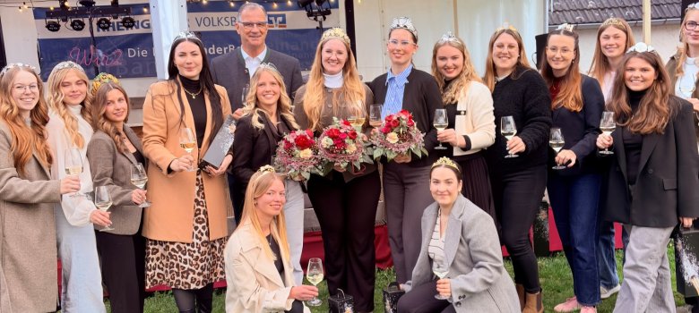 Fünfzehn junge Frauen stehen in einer Gruppe auf einer Wiese und lachen in die Kamera. Ein Mann ist dabei und trägt eine Brille und einen Anzug mit Hemd. Die Frauen tragen Jacken und überwiegend Hosen. Sie halten jede ein befülltes Weinglas in der Hand und haben Kronen auf dem Kopf. Zwei Frau Knien vor der Gruppe- Drei Frauen haben jeweils einen Blumenstrauß in der Hand. Im Hintergrund sind weiße Pavillons mit einer Lichterkette und Scheinwerfern aufgebaut.