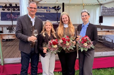 Drei junge Frauen stehen neben einem Mann mit weißem Hemd, JEans und Trachtenjacke. Er trägt eine Brille. Zwei der Frauen tragen ebenso eine Brille und alle Frauen haben Kronen im Haar und tragen Hosen und Jacken. Sie halten Blumensträuße u.a. mit roten Rosen in den Hänsen. Alle vier Personen haben ein gefülltes Weinglas in der Hand.