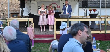 Auf einer Bühne stehen zwei Bistrotische mit grauen Hussen. An dem einen Tisch steht ein Mann mit blauem Anzug und weißem Hemd und Brille, der ein Mikrofon hält. Daneben stehen zwei Frauen im Dirndl mit Weingläsern in der Hand und goldener Krone in den blonden Haaren. Über der Bühne ist ein weißes Zeltdach. Im Vordergrund sitzen Menschen an Bierzeltgarnituren.