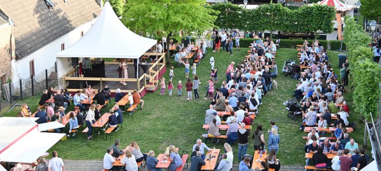Blick auf eine Wiese, auf der eine Bühne mit einem weißem Dachzelt ist und viele Bierzeltgarnituren stehen, auf denen Menschen sitzen. Links im Vordergrund ist ein geöffneter Weinstand  zu sehen. Rechts von der Bühne ist ein grüner Laubbaum. An zwei Seiten des Festplatzes sind Reihen von schmalen Laubbäumen.