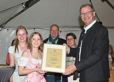 Bürgermeister Steinmacher im schwarzen Anzug und die neuen Weinprinzessin im rosa-weiss- farbenem Dirndl halten eine gerahmte Urlunde in der Hand. Links davon steht die Weinkönigin in beige-weißem Dirndl. Im Hintergrund steht eine weitere Frau und ein Mann. 