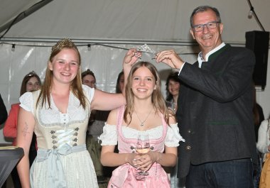 Bürgermeister Steinmacher im schwarzen Anzug und die Weinkönigin in beige-weißem Dirndl halten eine Krone über den Kopf der neuen Weinprinzessin im rosa-weiss- farbenem Dirndl  