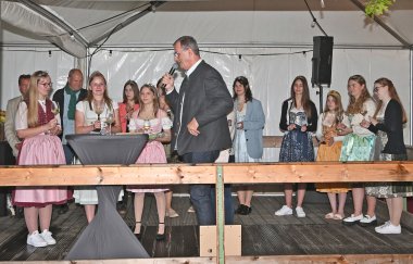 Auf einer Bühne aus Holz, die mit einer weißen Plane überdacht ist, stehen mehrere junge Frauen in Dirndeln und ein Mann im Anzug läuft mit Mikrofon in der Hand vor ihnen her. 