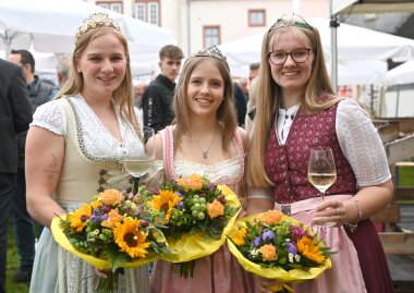Drei junge Frauen mit langen blonden Haaren im Dirndl und jeweils einem Blumenstrauß mit Sonnenblumen und Trauben in der Hand.