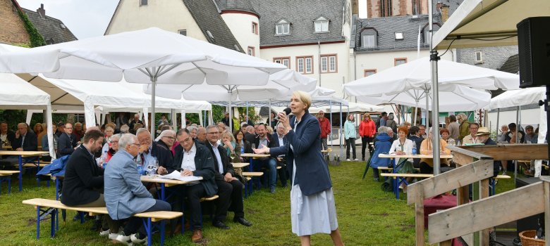 Man sieht eine Rasenfläche auf der mehrere Bierzeltgarnituren stehen, auf denen Menschen unter weiß, aufgespannten, großen Schirmen sitzen. Eine Frau steht vor einer Bühne und sinkt in ein Mkrofon, welches sie in der Hand hält. Im Hintergrund ist eine Kirche und ein beiges Haus zu sehen. 