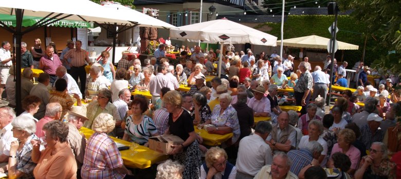 An mehreren Bierzeltgarnituren mit gelben Plastiktischdecken, sitzen mehrere Personen unter weißen Sonnenschirmen. Im Hintergrund ist ein Fachwerkhaus und ein Haus, das mit grünen Blättern bewachsen ist und rote Geranien aud den Fensterbänken hat.