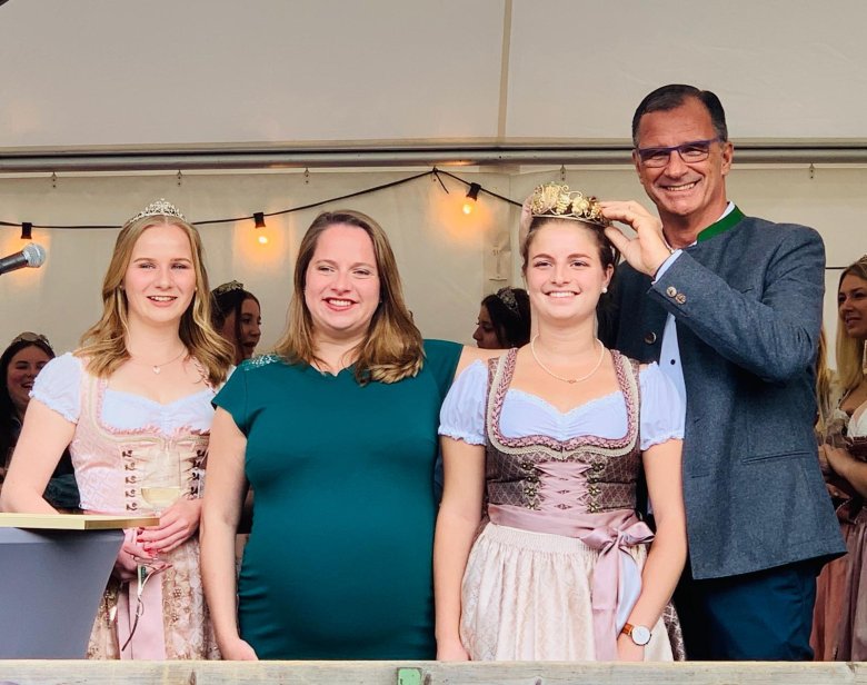 Die Weinkönigin und Weinprinzessin tragen eine weiße Bluse und rosa-beige Dirndl mit Schürze. In der Mitte steht die schwangere, ehemalige Weinkönigin in einem türkisem Kleid. Bürgermeister Winfried Steinmacher, im blauem Anzug, hält das goldene Diadem und setzt es der künftigen Weinkönigin auf den Kopf.  
