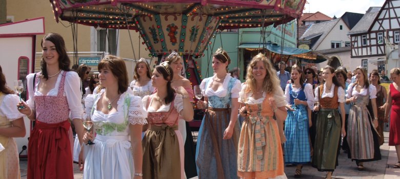 Mehrere Frauen in Dirndls mit Weingläsern in der Hand und Kronen auf dem Kopf, laufen eine Straße entlang. Auf der Straße steht ein Kettenkarussell für Kinder und eine Verkaufsbude in türkis. Es sind Häuser und Fachwerkhäuser zu sehen. 