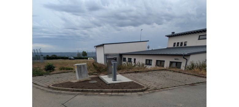 Je zwei gepflasterte Parkplätze, im rechten Winkel und dazwischen auf einem dreieckigem Erdstück steht ein Stromkasten und zwei silberne Säulen. Im Hintergrund sind beige Gebäude mit Fenstern.