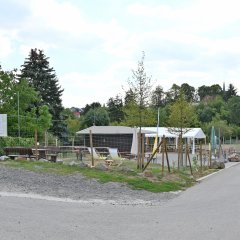 Asphaltierte landwirtschaftliche Straße und Drahtzaun mit Holzpfählen. Innen der Weinstand aus Holz, ein weißes, rechteckiges Pavillon,  weiße und grüne zusammengefaltete Sonnenschirme, Sonnenstühle, Bänke, zwei große Schilder und im Hintergrund Laubbäume.