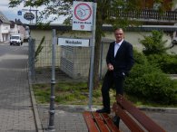 Mit-Fahr-Bank Kiedrich Hinter einer braunen Bank steht Bürgermeister Winfried Steinmacher mit weißem Hemd und schwarzem Anzug. Links davon ist ein weißes, rechteckiges Schild mit einem rotem Kreis , in dem auf einem weißem Kreis eine Hand mit Daumen hoch gezeichnet ist. Unter dem Schild hängt ein weiteres rechteckiges, weißes Schild, auf dem "Weisbaden" in schwarzen Buchstaben geschrieben ist.