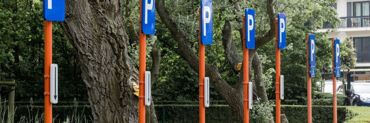 Im Vordergrund ist eine Kreisförmige Hecke. Im Kreis stehen alte große Laubbäume. Zwischen den Bäumen und der Hecke stehen sieben Schilder, auf orangenen Stangen. Oben sind blaue rechteckige Schilder, auf dneen ein weißes "P" steht. An den Stangen sind weiße, rechteckige Schilder, auf denen ein schwarzer Pfeil nach unten zeigt.