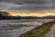 Am Flussufer ist ein Weg mit Steinen und ein Grasrand. Auf dem Fluss fährt Transportschiff mit Waren. Im Hintergrund sind verschiedene unbelaubte schwarze Bäume. Der Himmel ist mit dunklen Wolken versheen und am Horizont ist ein heller Streifen ohne Wolken.