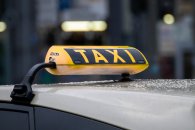 Das schwarz-gelbe Taxirechteck in dreiecksform auf dem Dach des beigen Taxis. Im Hintergrund sieht man verschwommen ein weißes Vordach, Hecken und eine Straße.