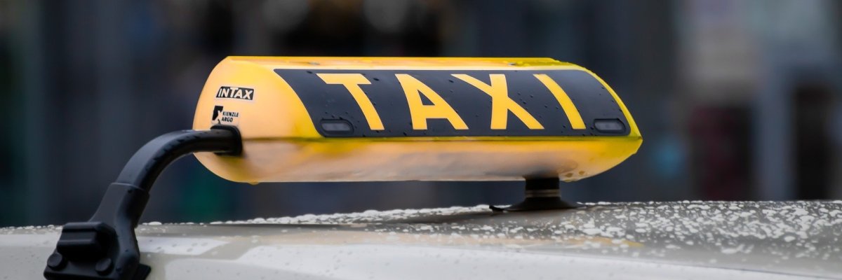 Das schwarz-gelbe Taxirechteck in dreiecksform auf dem Dach des beigen Taxis. Im Hintergrund sieht man verschwommen ein weißes Vordach, Hecken und eine Straße.