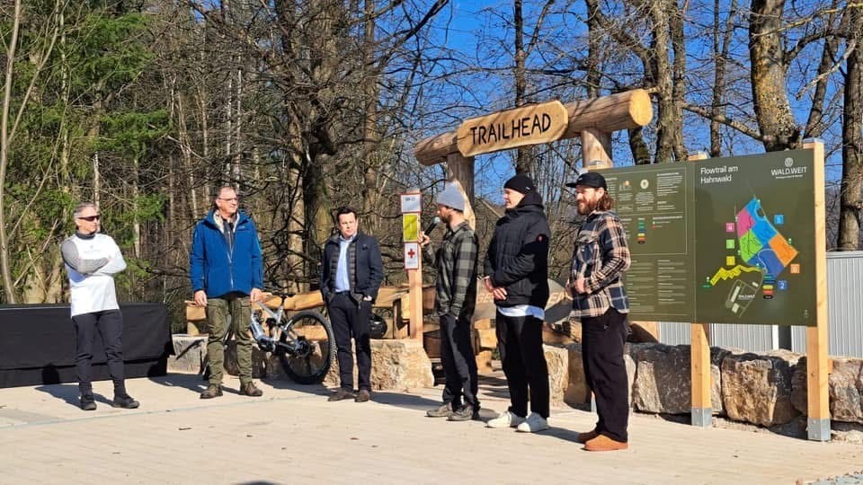 Man sieht Bürgermeister Steinmacher, sowie fünf Männer und ein Mountainbike vor dem Eingang zur Mountainbike-Strecke, der aus großen Holzstämmen zusammen gebaut wurde. Oben hängt ein Holzschild mit "Trailhead". Rechts davon ist eine Hinweistafel u.a. mit den Routen und Öffnungszeiten an drei großen Holzpfählen angebracht. Im Hintergrund ist der Wald.