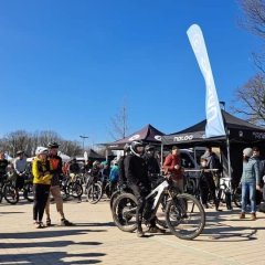 Auf einem asphaltierten Parkplatz stehen mehrere Personen und viele Mountainbikes. Es sind mehrere Pavillons und mehrere Dropflags aufgestellt. Im Hintergrund sind Bäume und ein blauer Himmel. 