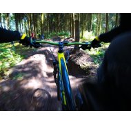 Auf einem schmalen, unebenem Waldweg, fahren mehrere Mountainbikes. Im Vordergrund sieht man das Vorderrad, das grüne Lenkrad und die gelbe Achse. Der Fahrer trägt schwarze Handschuhe und eine schwarze Jacke mit einem gelben Bund. Um den Weg herum sind viele Bäume.