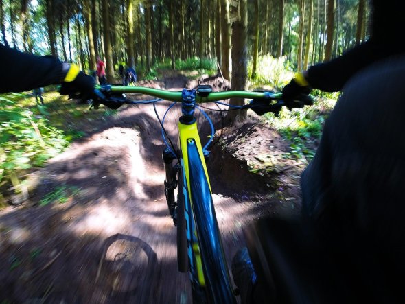 Auf einem schmalen, unebenem Waldweg, fahren mehrere Mountainbikes. Im Vordergrund sieht man das Vorderrad, das grüne Lenkrad und die gelbe Achse. Der Fahrer trägt schwarze Handschuhe und eine schwarze Jacke mit einem gelben Bund. Um den Weg herum sind viele Bäume.
