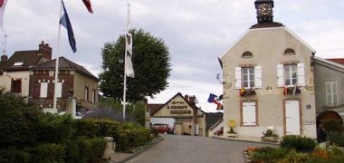 Eine Straße läuft auf mehrere beige Häuser zu. Links und rechts sind Beete mit Sträuchern und Hecken, die von Steinen umrandet sind. Auf dem Rathaus ist ein Schieferturm, auf dem sich eine Uhr mit goldenen Zeigern und Strichen befindet. Links im Bild sind drei Fahnenmaste, an denen jeweils zwei Fahnen wehen.
