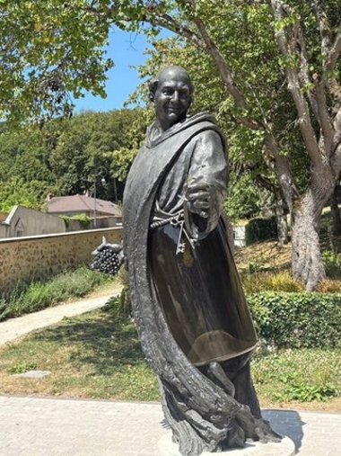 Eine Statue aus grauem Metall zeigt einen Mönch mit einem Schlüsselbund an einer Kordel, die er als Gürtel trägt. Er hält Trauben in der Hand. Im Hintergrund ist ein hell gepflasterter Weg und eine Mauer und Bäume.