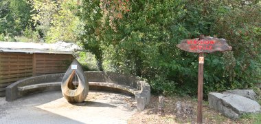 Tropfenförmige Skulptur, aus der oben Wasser aus der Virchow-Quelle fliesst mit einer halbkreisförmigen Steinbank und Bäumen und einem Hinweisschild aus Holz mit roter Schrift