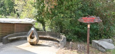 Virchow-Quelle Tropfenförmige Skulptur, aus der oben Wasser aus der Virchow-Quelle fliesst mit einer halbkreisförmigen Steinbank und Bäumen und einem Hinweisschild aus Holz mit roter Schrift