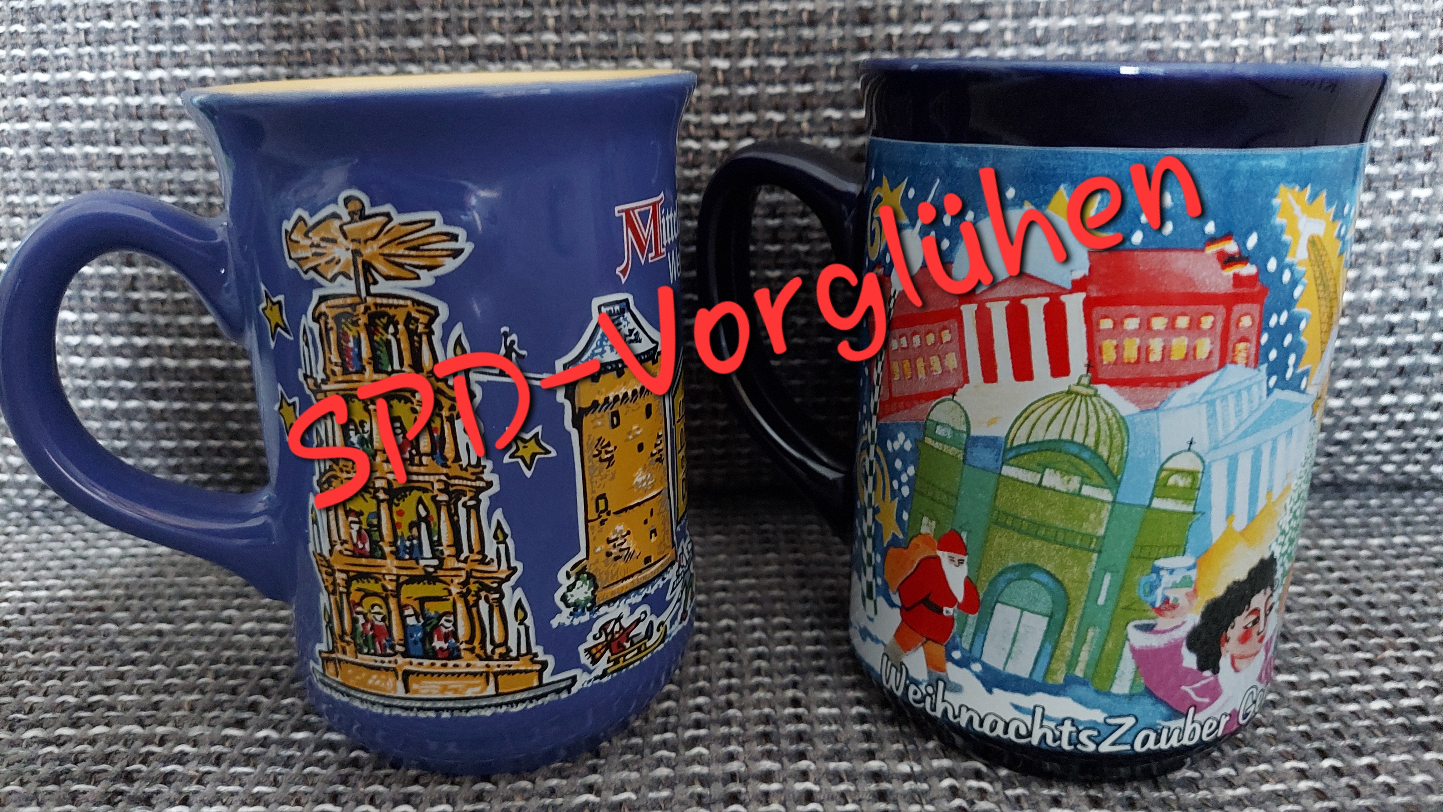 Auf einem grau-weiß-schwarz gemustertem Stoff stehen zwei blaue Glühweintassen mit Weihnachtsmotiven, u.a. eine Holzpyramide, ein Nikolaus und verschiedene Gebäude.
