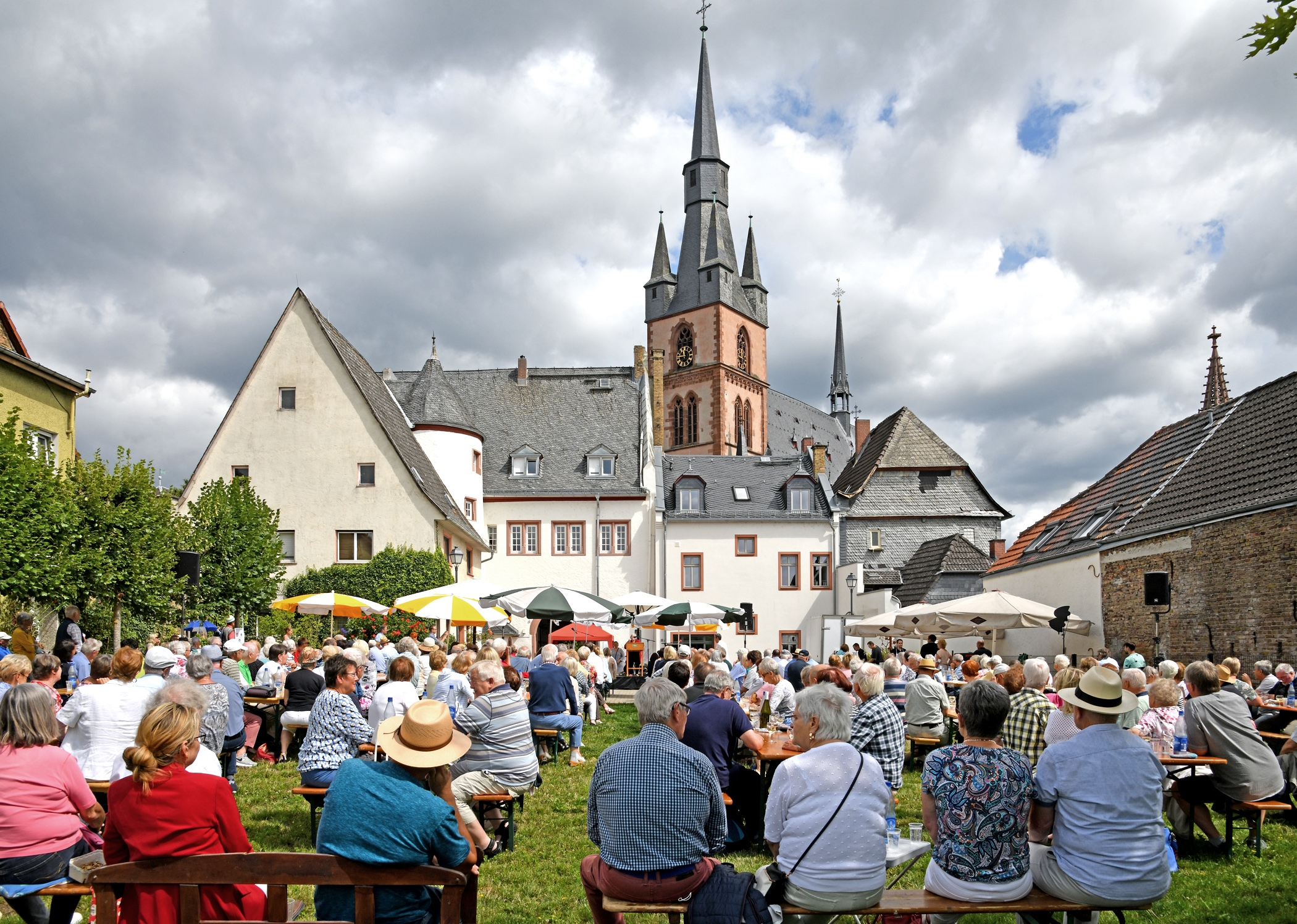 Auf der Wiese des Josef-Staab-Platzes sitzen auf Bierzeltgarnituren Frauen und Männer, zum Teil unter aufgespannten Sonnenschirmen. Im Hintergrund das weiße Rathaus mit den grauen Dachziegeln und den roten Fensterrahmen und dahinter die Basilika minor vor einem bewölktem Himmel.