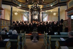 Ein gemischter Chor, 46 Personen, in schwarz gekleidet und mit schwarzen Gesangsbüchern in der Hand, steht im Altarraum. Hinter ihnen ein Hochaltar, dahinter zwei große Sprossenfenster und auf einer Empore die Orgel. Vorn sind Kirchenbänke mit Zuhörern zu sehen. Die Kirche ist in hell- und dunkelgrün und gold gehalten.